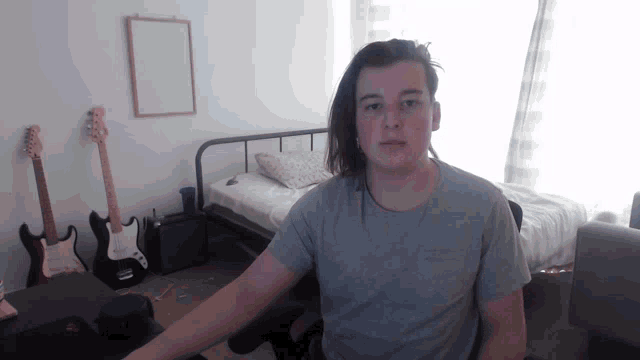 a man in a grey shirt is sitting in front of a bed and guitars
