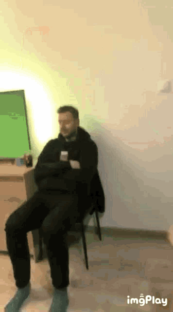 a man is sitting in a chair with his arms crossed and a green screen behind him .