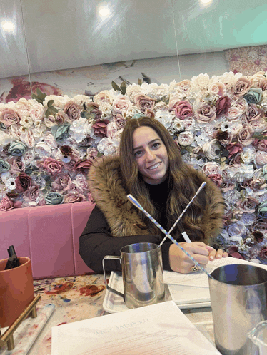 a woman sits at a table in front of a wall of pink flowers