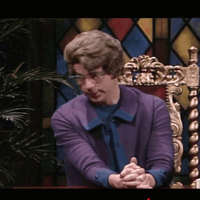 a man in a purple sweater sits in a chair with his hands folded