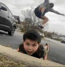 a boy is crawling on the sidewalk next to a car while a man is jumping in the air .