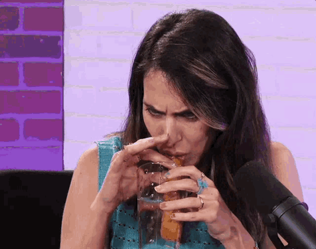 a woman is drinking from a glass while sitting in front of a microphone in front of a brick wall .