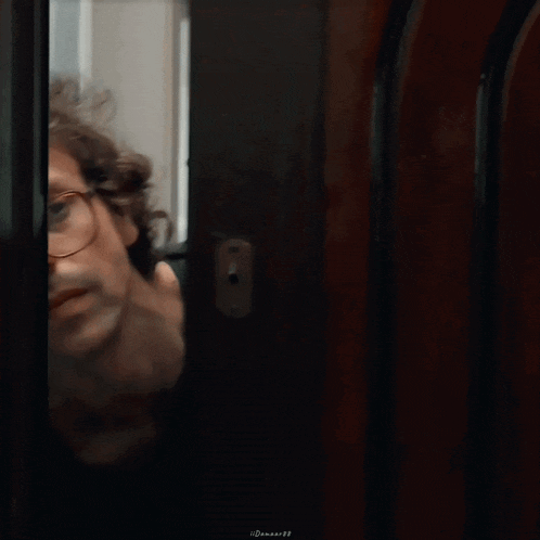 a man with curly hair wearing glasses is standing in front of a wooden door