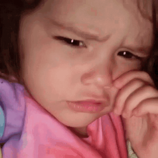 a little girl in a pink shirt is crying and covering her nose with her hand .