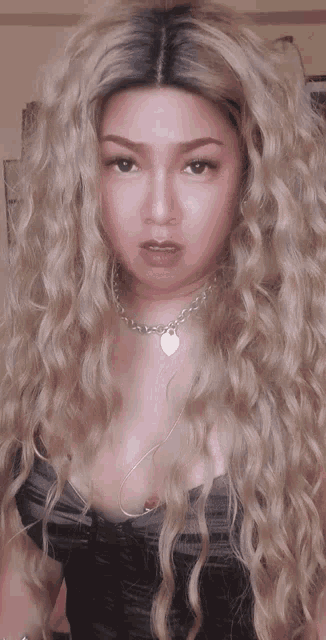 a woman with long blonde curly hair is wearing a necklace and a black top