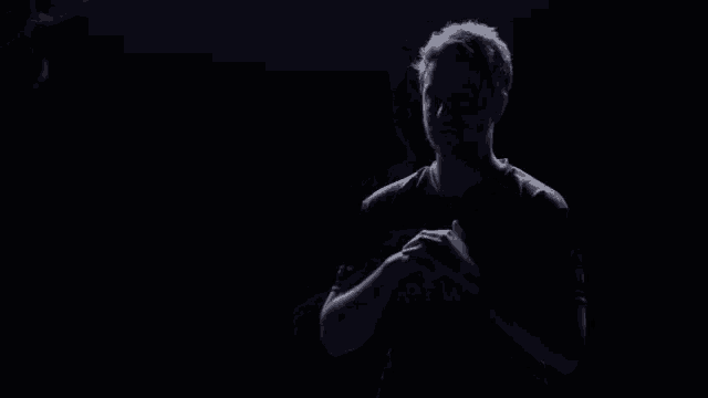 a man in a black shirt is standing in a dark room with his hands folded .