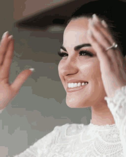 a woman in a white lace top is smiling with her hands in front of her face