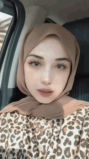 a woman wearing a leopard print shirt and hijab is sitting in a car