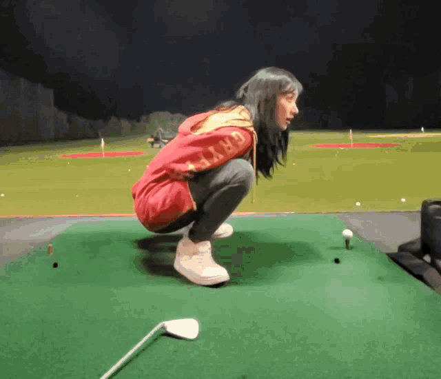 a woman wearing a red jacket with the word gryffindor on the sleeve squats on a golf course