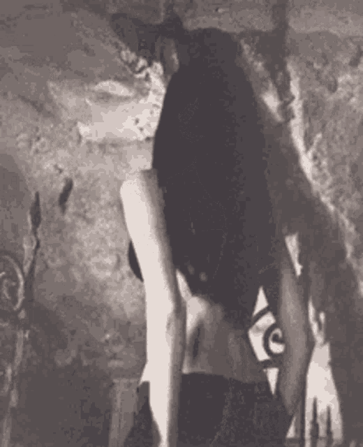 a black and white photo of a woman with long black hair standing in front of a wall .