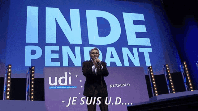 a man stands on a stage in front of a large blue sign that says inde pendant