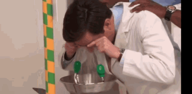 a man in a lab coat is washing his face in a sink with a green faucet .
