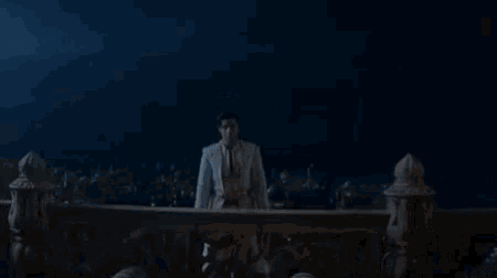 a man in a white coat is standing on top of a balcony overlooking a city at night .