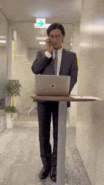 a man in a suit and tie is talking on a cell phone while standing next to a laptop on a table .