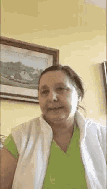 a woman in a green shirt and a white vest is smiling in front of a painting .