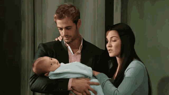 a man in a suit holds a baby while a woman looks on