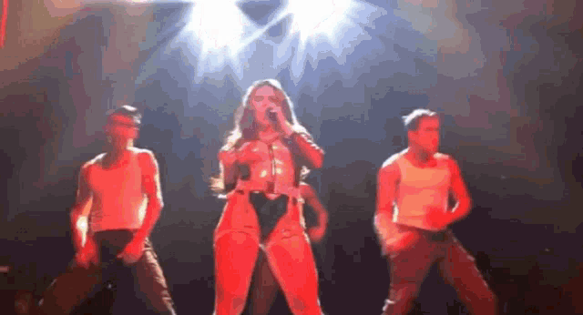 a woman is singing into a microphone while two men dance behind her on a stage
