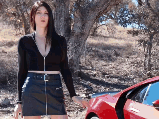 a woman in a black crop top and black skirt is standing next to a red car