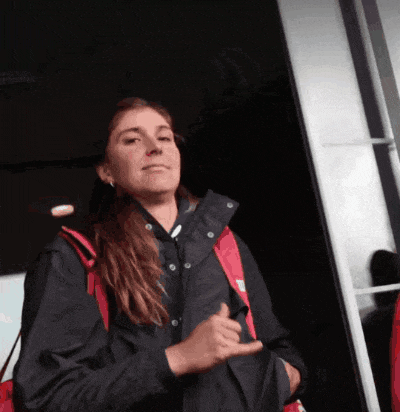a woman wearing a black jacket and a red backpack is making a hand sign