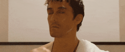 a shirtless man with a towel around his neck is sitting in front of a white wall with his eyes closed .