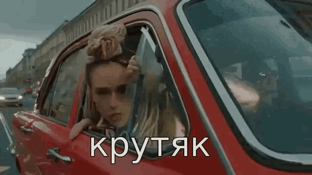 a woman is sticking her head out of the window of a red car while driving down a street .
