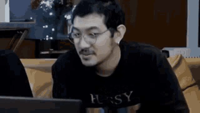 a man wearing glasses and a black shirt is sitting on a couch looking at a laptop computer .