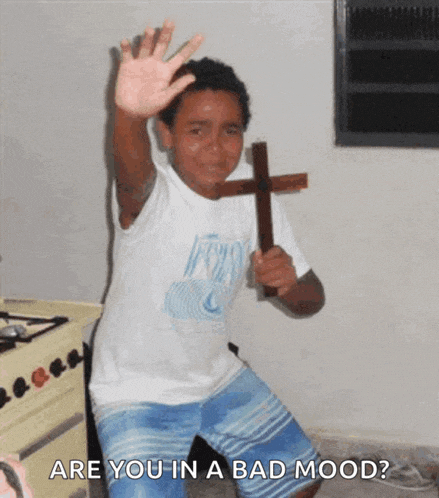 a young boy is holding a wooden cross and waving at the camera with the words are you in a bad mood below him