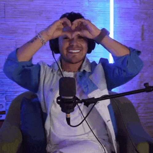 a man wearing headphones is sitting in front of a microphone making a heart shape with his hands .