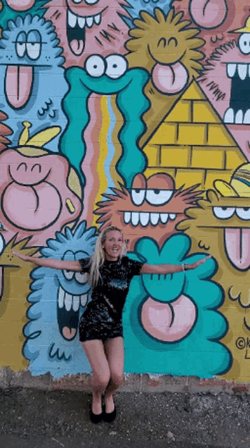 a woman in a black dress stands in front of a wall with cartoon faces on it
