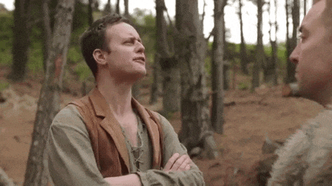 a man is standing in the woods with his arms crossed talking to another man .