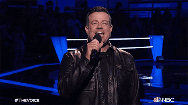 a man in a leather jacket singing into a microphone with the nbc logo behind him