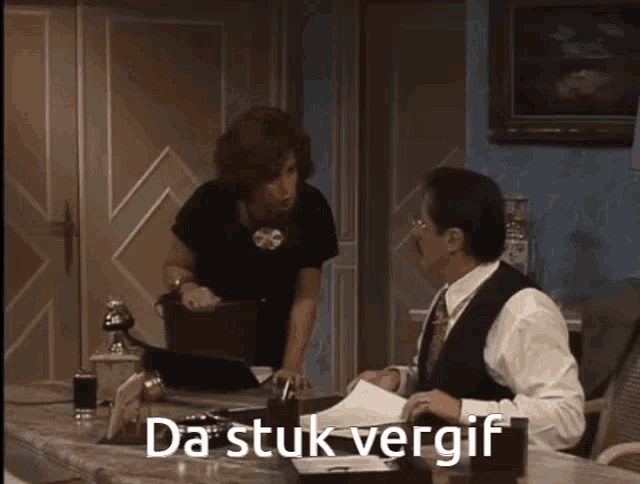 a man sitting at a desk with a woman standing behind him with the words da stuk vergif written on the screen