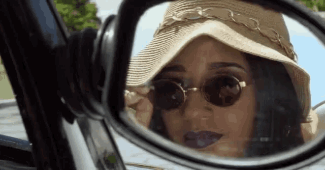 a woman wearing a hat and sunglasses is looking at herself in a car rear view mirror .