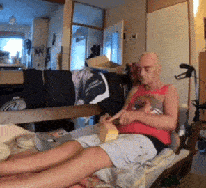 a man in a red tank top sits on a bed with his legs crossed