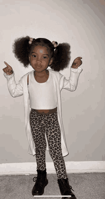 a little girl wearing leopard print pants and a white cardigan is pointing up