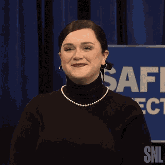 a woman wearing a black turtleneck and pearls stands in front of a sign that says safi