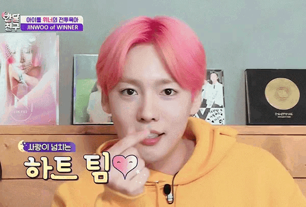 a young man with pink hair is wearing a yellow hoodie and making a heart shape with his finger