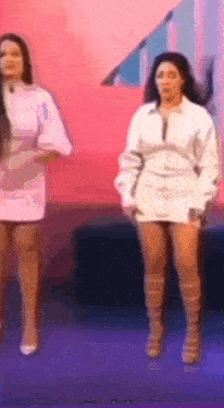 two women are standing next to each other on a stage . one is wearing a white dress and the other is wearing brown boots .
