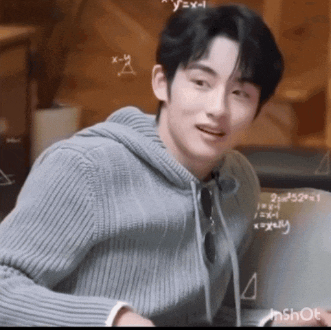a young man in a gray sweater is sitting in front of a chalkboard with mathematical equations on it .