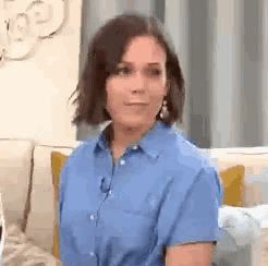 a woman in a blue shirt is sitting on a couch looking at the camera .