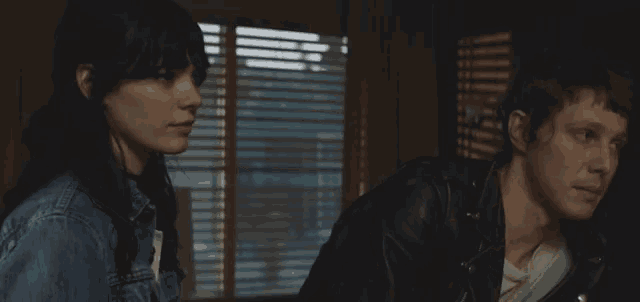 a man and a woman looking at each other in front of a window with blinds