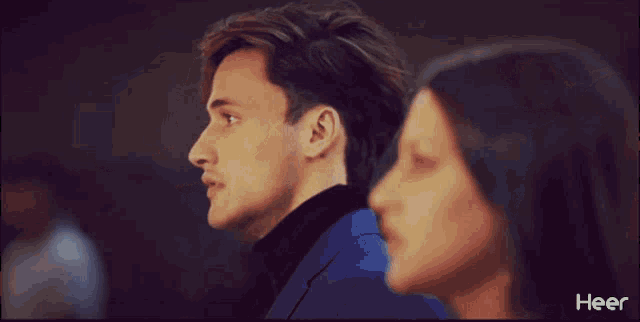a man in a blue suit and black turtleneck looks at a woman in a heer ad