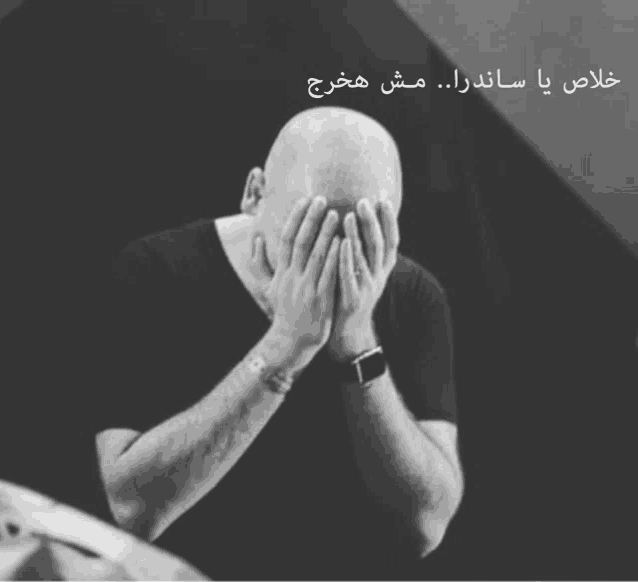 a bald man covering his face with his hands with arabic writing behind him