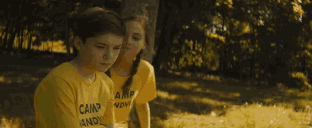 a boy and a girl are sitting under a tree wearing yellow camp shirts