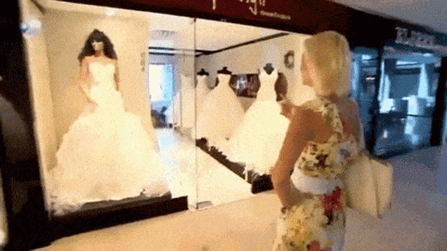 a woman in a floral dress looks at a wedding dress in a store window