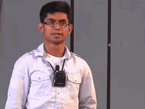 a man wearing glasses and a white shirt is standing in front of a wall and says `` good '' .