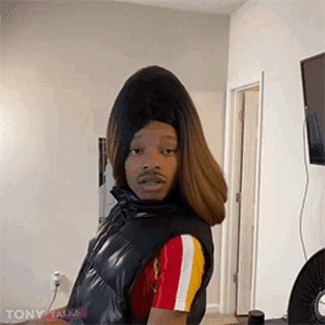 a man wearing a wig and a vest is standing in a living room in front of a fan .