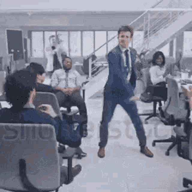 a man in a suit and tie is dancing in an office with other people sitting in chairs .