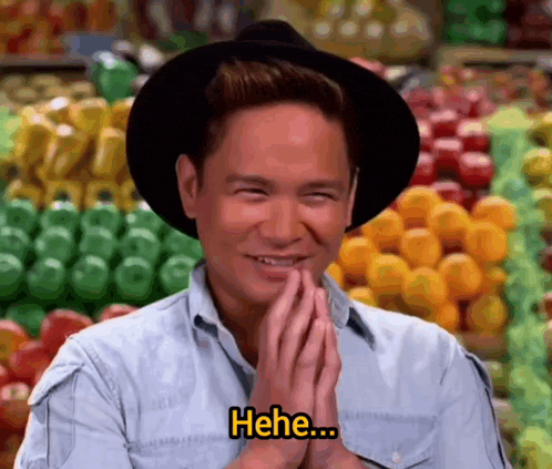 a man wearing a hat and a blue shirt is smiling and saying " hee " in front of fruits and vegetables