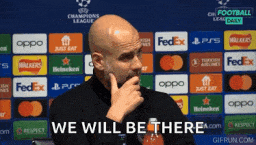 a bald man with a beard is standing in front of a row of advertisements and says we will be there .
